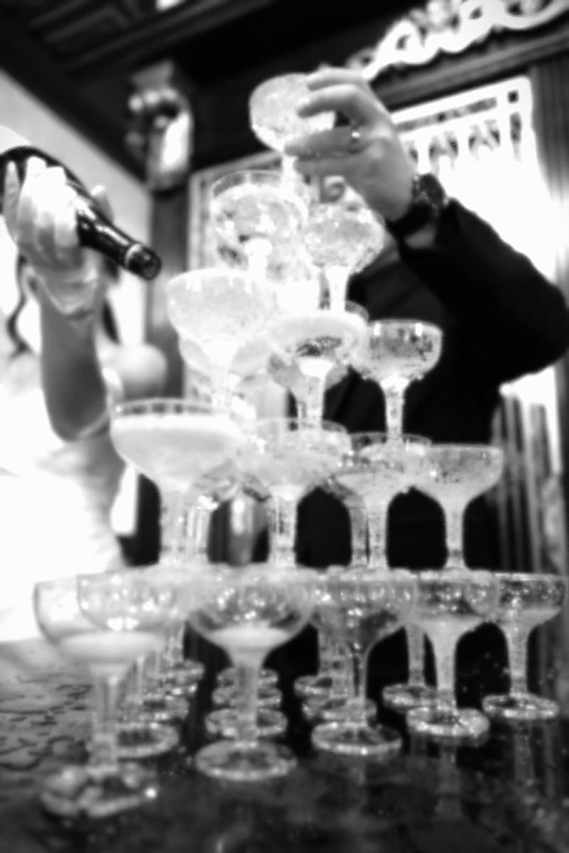 A bride pouring champaign into a tower of glasses, as her groom makes a toast.